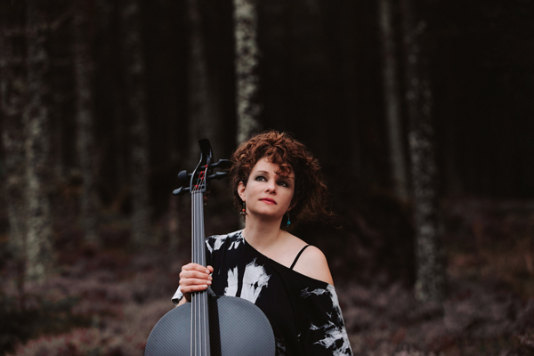 Beth Porter image. Beth is wearing a black and white top, she is stood in front of a forest with her black cello in her hand. 