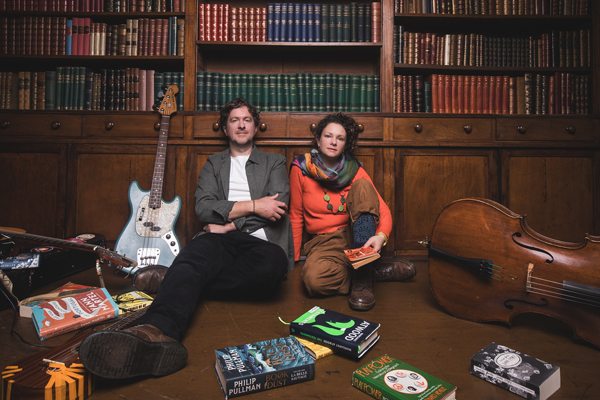 The Bookshop Band image. The duo are sat on the floor of an old library. There are dark cabinets behind them filled with books. There are some books scattered around them on the floor, as well as some instruments too. 