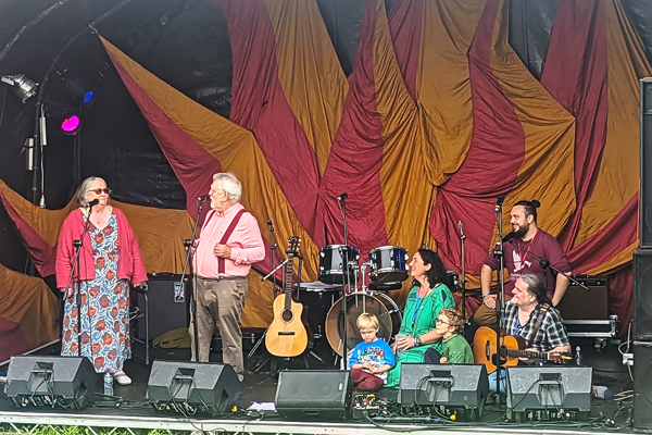 Burke Family Band image. The family are on the Fire Stage at Purbeck Valley Folk Festival. There is a bright red and orange backdrop for the stage. Paul & Sheila Burke are stood in front of microphones on the left hand side of the stage, and 5 other family members (2 of their children, and 3 grandsons) are sat down on the right hand side of the stage.