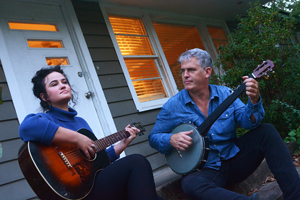 Dirk & Amelia Powell image. Amelia and Dirk are sat down on a step outside of a house. Amelia is playing her guitar, and Dirk is playing his banjo.