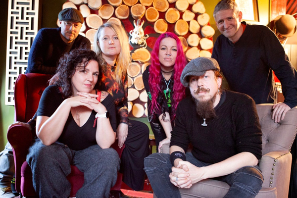 High Shelf Remedy image. 6 band members are posing for a photo. Two are in the foreground sat forward in big armchairs, and 4 are stood behind. 3 males, 3 females. 