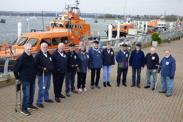 Wareham Whalers image. 11 members of Wareham Whalers all stood outdoors, next to a docked orange lifeboat. They are all wearing matching blue jumpers that have the Wareham Whalers logo on it. 