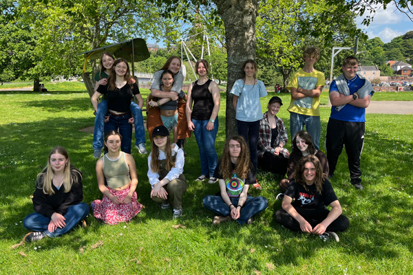 Devon Youth Folk Ensemble image. 15 young musicians are photographed on a green field in front of a tree (they are in the shade). Half of them are sat down on the grass, and the other half are stood behind them. They are all smiling and two people are giving piggy backs!