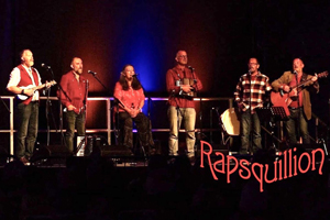 Rapsquillion image. Several members on stage all wearing a different type / style of red top, singing. 