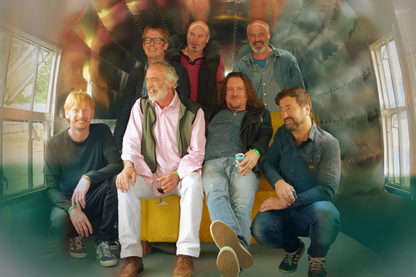 7 members of Kíla are in the image, all with big smiles. They are inside a silver bullet style caravan. 2 of them are sat on a yellow sofa and the rest are either crouched to the side or stood behind the sofa. A few of them have drinks in hand and the silver caravan roof above them is creating a colourful reflection. 