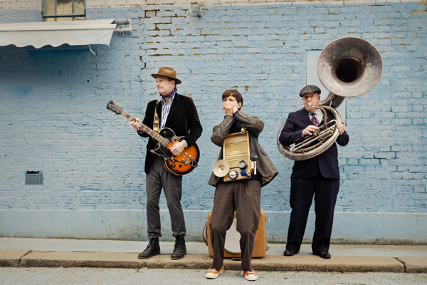 The Rigmarollers are stood in front of a blue brick wall outside. There are 3 of them and they are playing their instruments (harmonica, washboard, guitar, sousaphone)